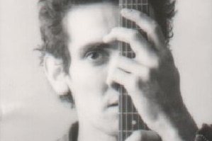 Black and white grainy photo of a young Paul Kelly with spiky dark hair holding the neck of a guitar in front of his face.