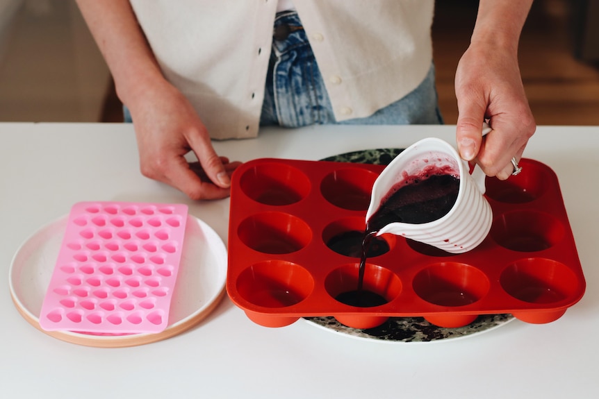 A pair of hands pour red liquid from a jug into silicone moulds on a bench.
