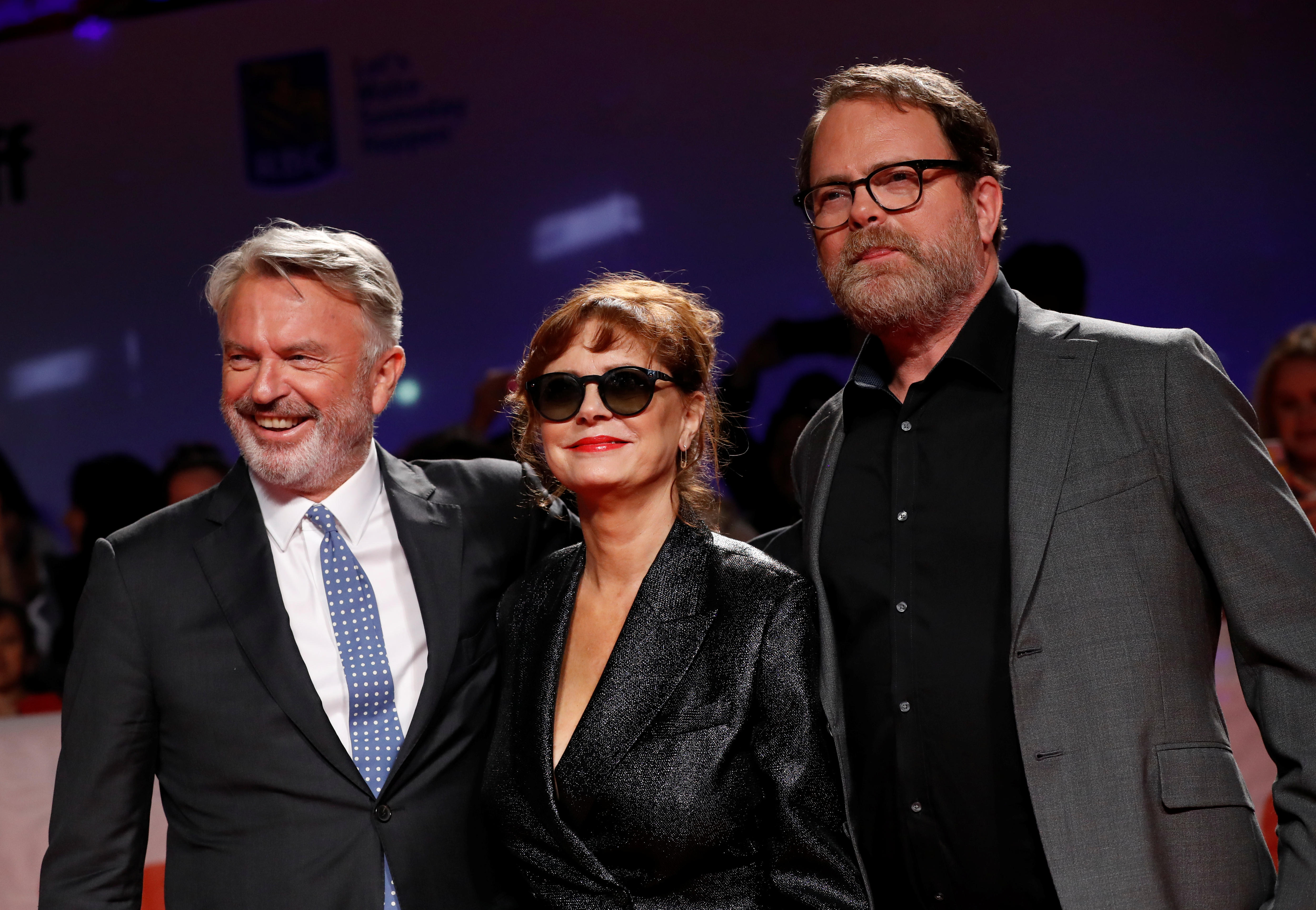 Sam Neill in a dark suit smiling next to Susan Sarandon in black sunglasses and a jacket, and Rainn Wilson.