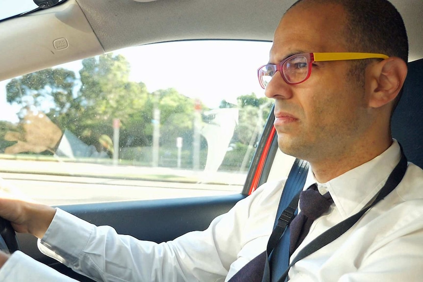A man behind the wheel of a car wearing glasses and a shirt and tie.