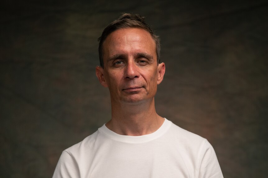 A man with short brown hair wearing a white tshirt looks at the camera. He sits in front of a dark brown background.