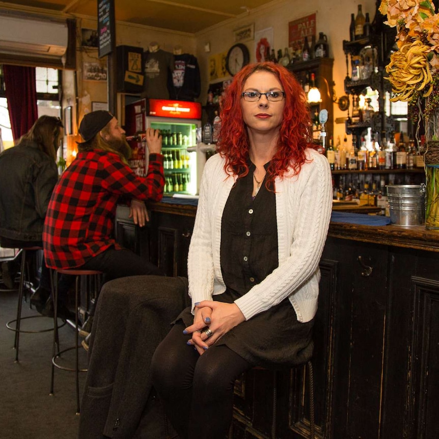 A woman sits at a bar, with drinkers in the background.