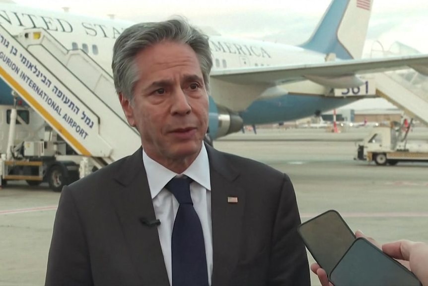 Antony Blinken wears a suit and stands on the tarmac infront of governmemt plane.