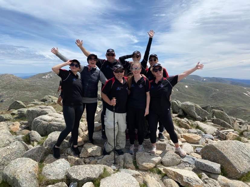 eight people in hiking clothing stand with their hands in the air facing the camera at the top of a mountains