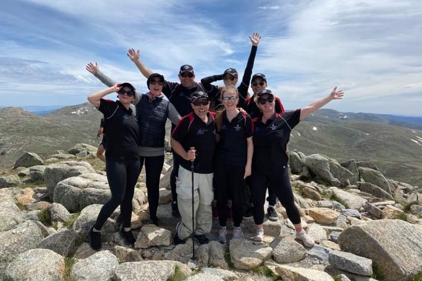eight people in hiking clothing stand with their hands in the air facing the camera at the top of a mountains
