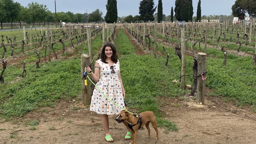 Eliza Berlage standing in front of some grape vines in a white dress with her dog Honey