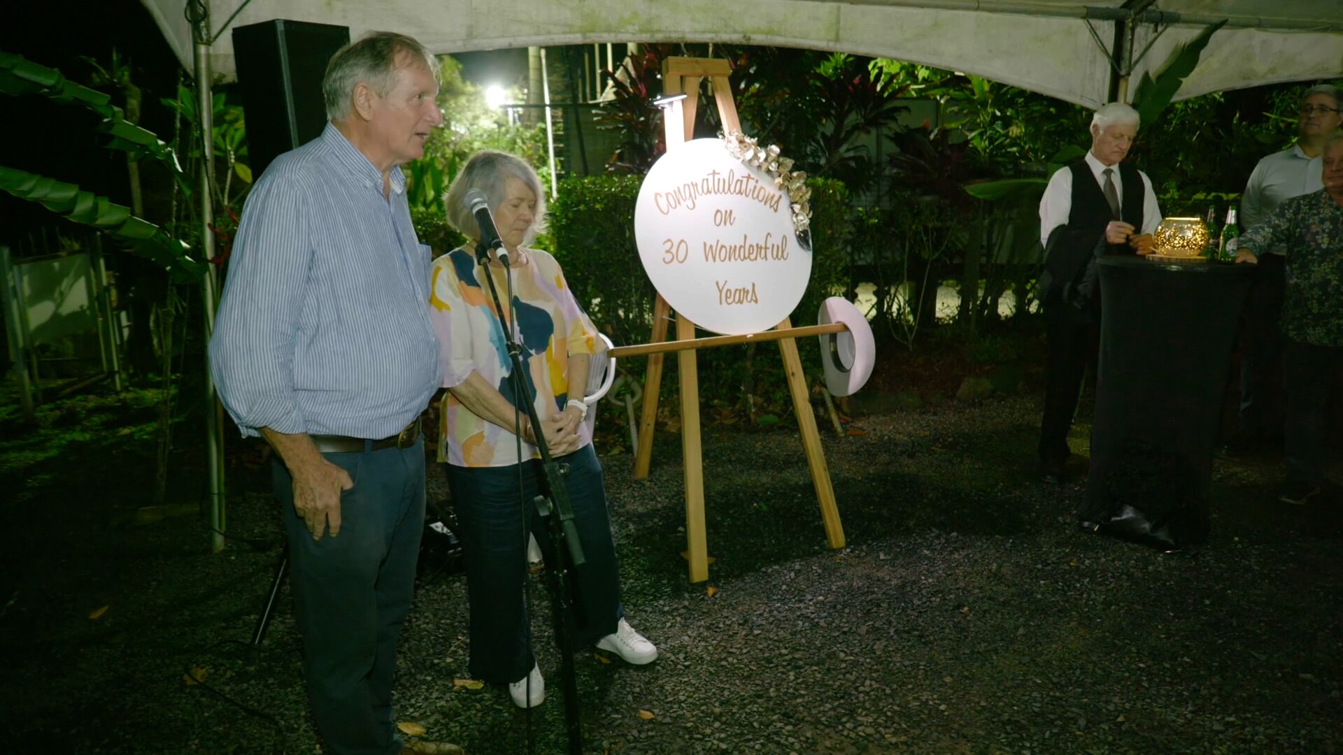 Dos personas mayores dan un discurso, cerca de un cartel que dice Felicitaciones por 30 maravillosos años.