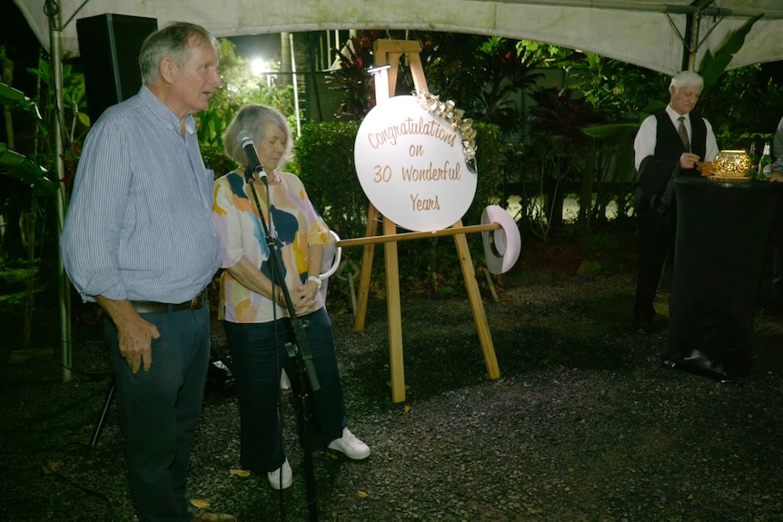 Two older people stand giving a speech, near a sign that says Congratulations on 30 wonderful years