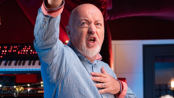 Bill Bailey stands with one hand raised, the other over his heart, singing joyfully in front of bank of keyboards