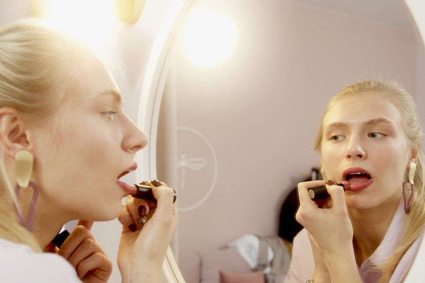 A blonde woman applies lipstick in a mirror. 