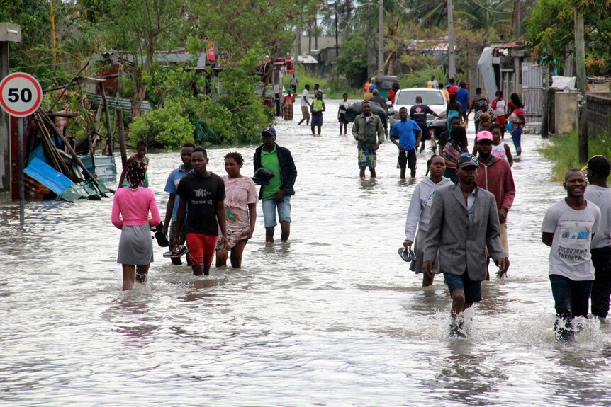 People walk down a flooded road with corrugated iron homes on either side.
