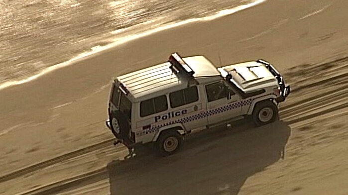 A police four-wheel drive drives down a beach, on its left waves hit the shore.