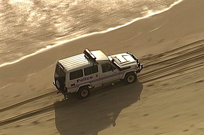 A police four-wheel drive drives down a beach, on its left waves hit the shore.