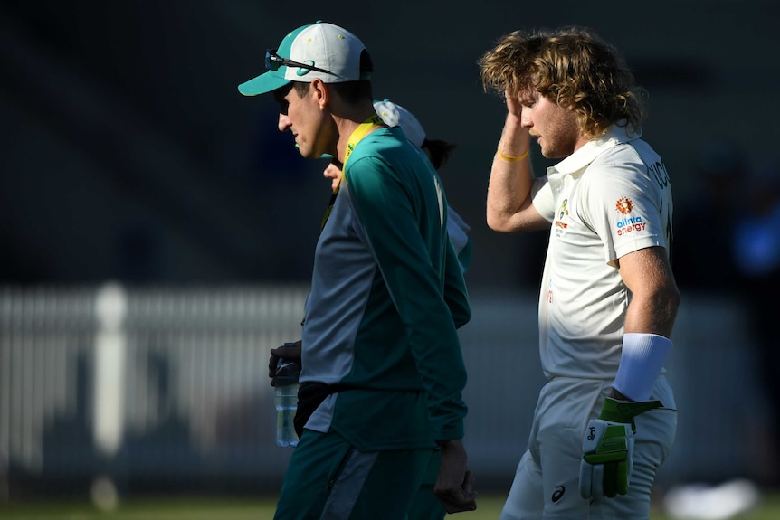 An Australian A male batter walks off the field holding his head after sustaining a concussion.