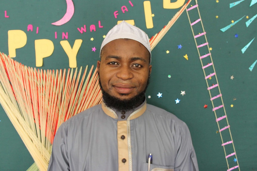 A South African man in traditional Islamic attire stands in front of a green wall of Eid festival celebrations.