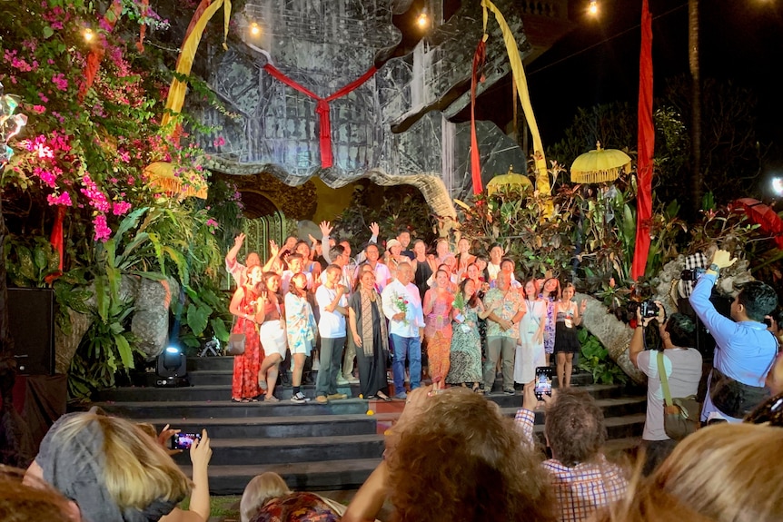 A crowd of people stand on stairs waving in front of an audience, surrounded by tropical plants