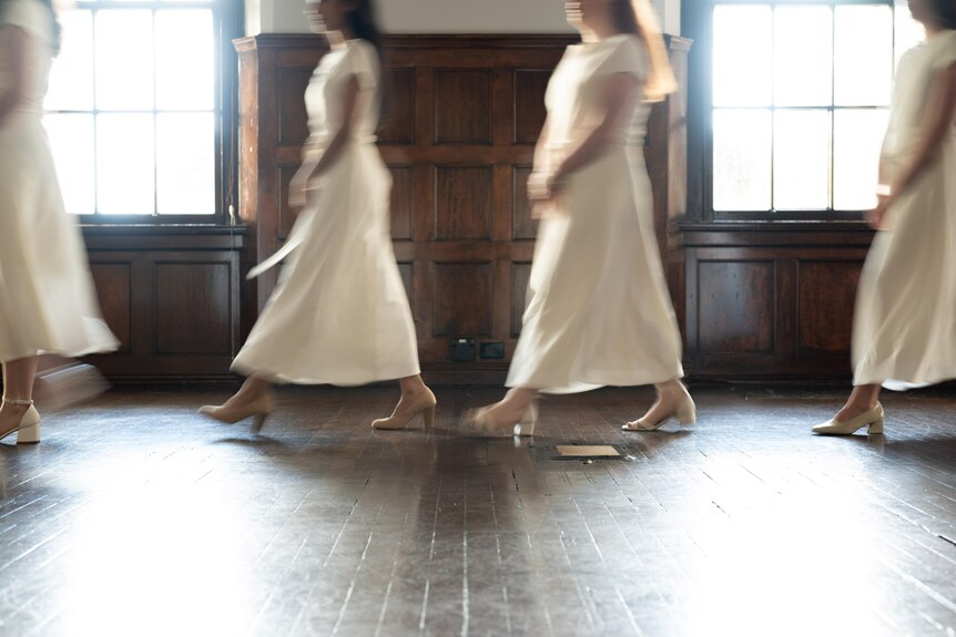 Four young women in matching white dresses walk in a line in a wood-paneled room with light-filled windows.