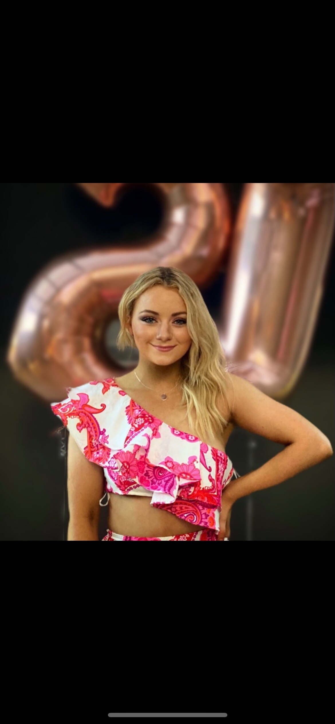 A blond woman smiling in a dress with pink balloons in the background
