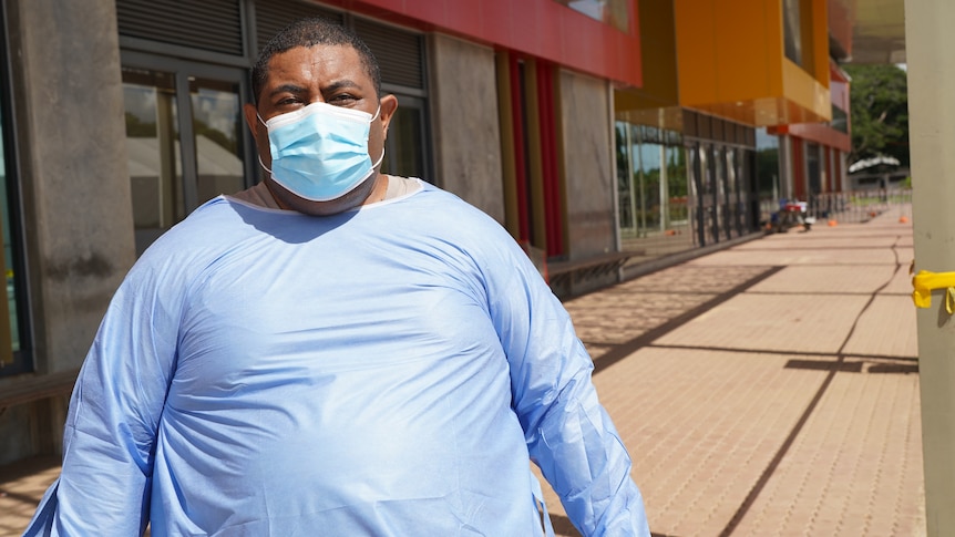 A man wearing dark blue scrubs and a mask stares at the camera /