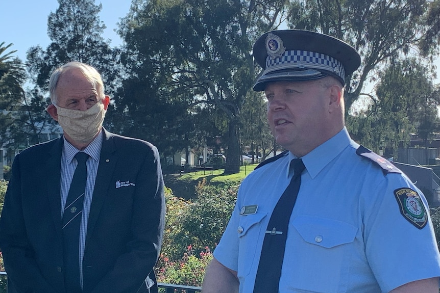 Man wearing navy jacket and yellow face mask stands next to a police officer in a blue shirt