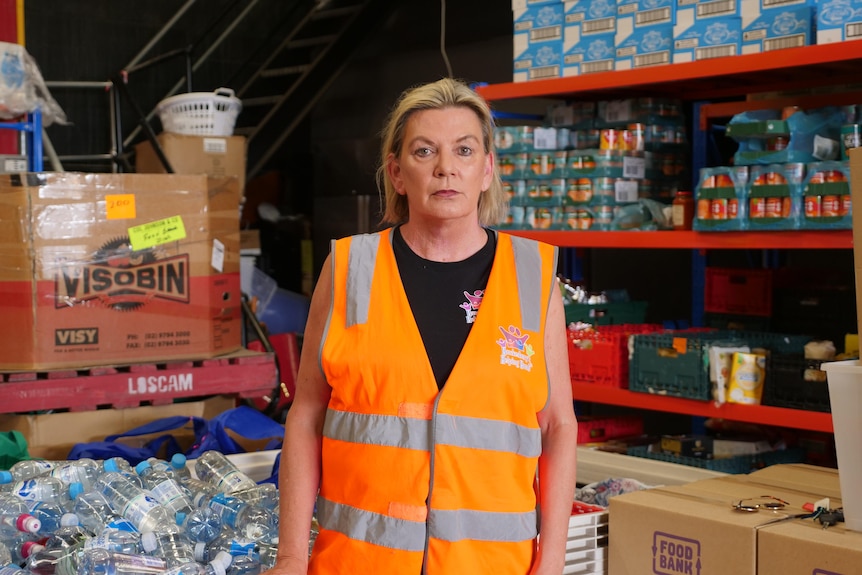 A woman looks neutrally standing among shelves of tins of food and water bottles