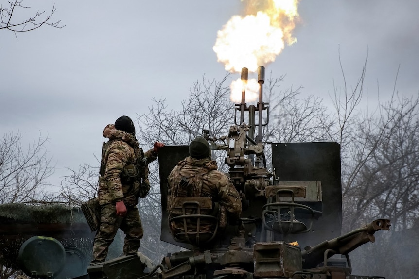 Two soldiers, with their backs to a camera, operating a large machine outdoors with flames coming out of it