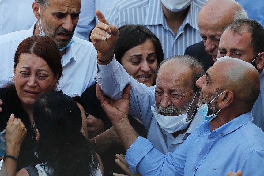 A group of mourners, some with face masks lowered, cry and comfort each other.