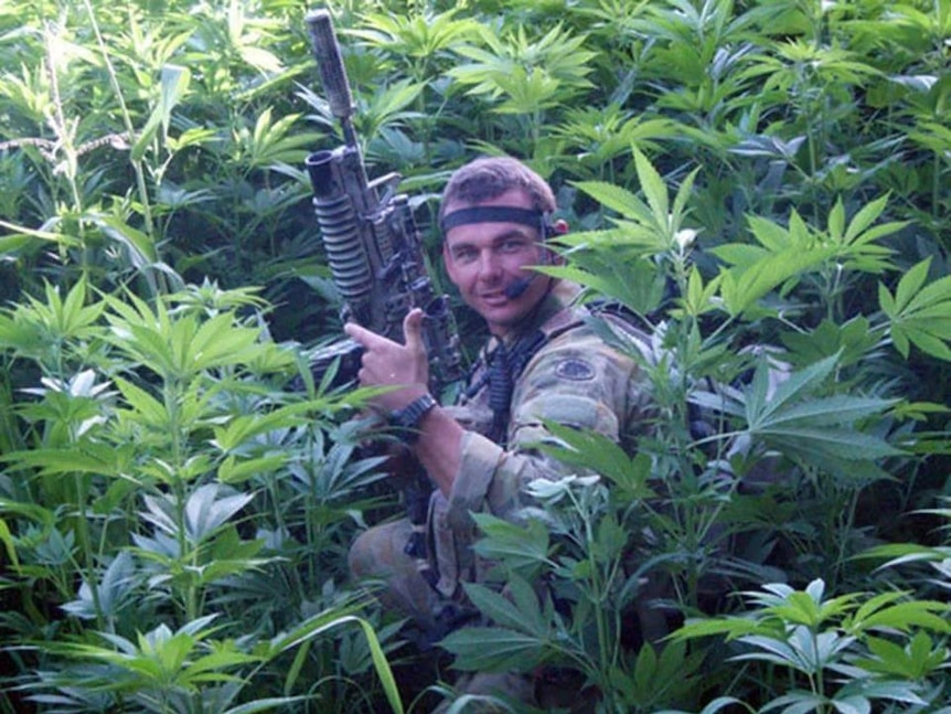 Commando Kevin Frost crouches among marijuana plants in Afghanistan holding a large gun and wearing camouflage gear.