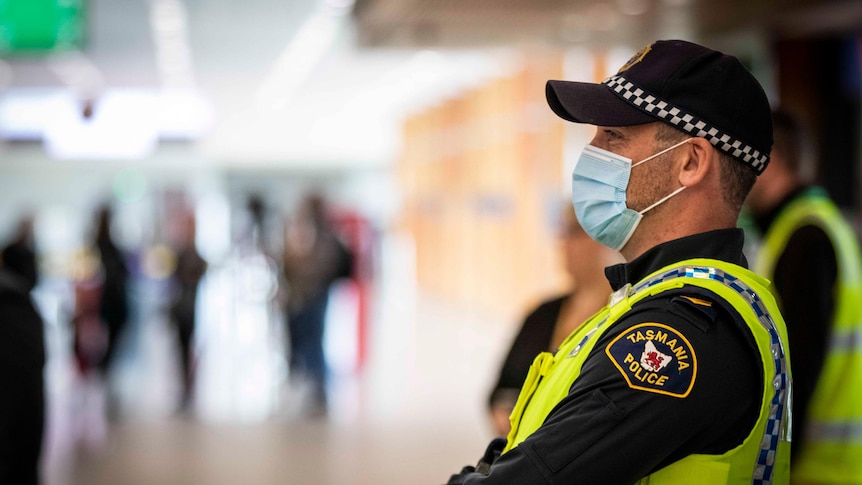 A male policeman wearing a mask at Hobart Airport, waiting in the arrivals hall.  He wears high visibility.