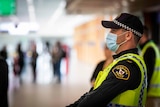 A male police officer in a mask at Hobart airport, waiting in the arrivals hall. He is wearing hi-vis.