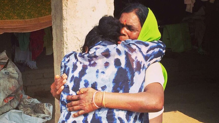 Two women embrace each other in an emotional hug. One is wearing a bright green sari, the other a blue and white top.