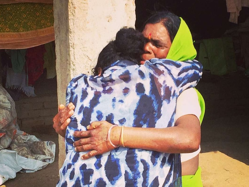 Two women embrace each other in an emotional hug. One is wearing a bright green sari, the other a blue and white top.
