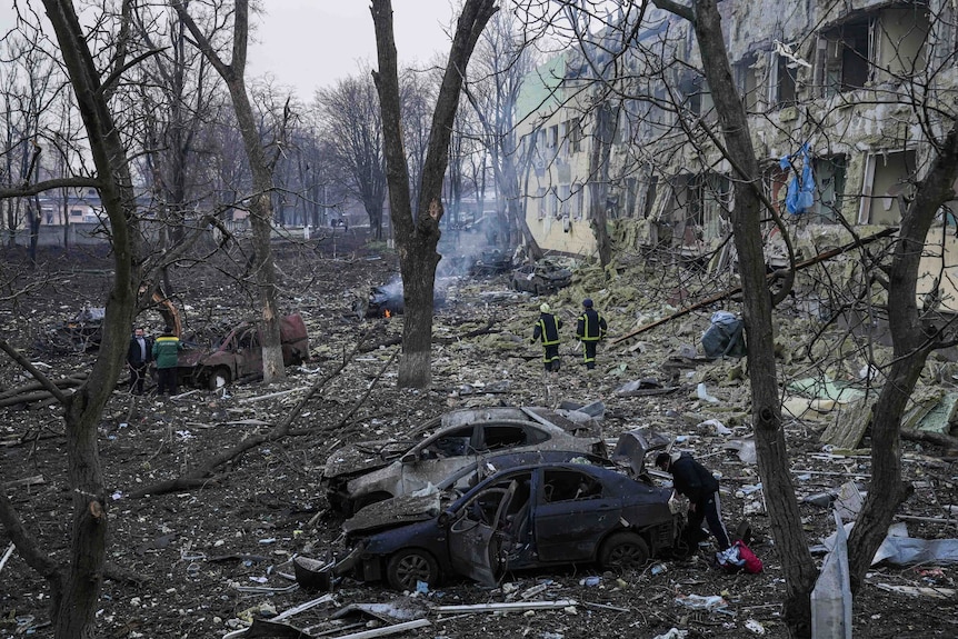 People stand among burnt-out cars, rubble and trees, outside a damaged hospital building.