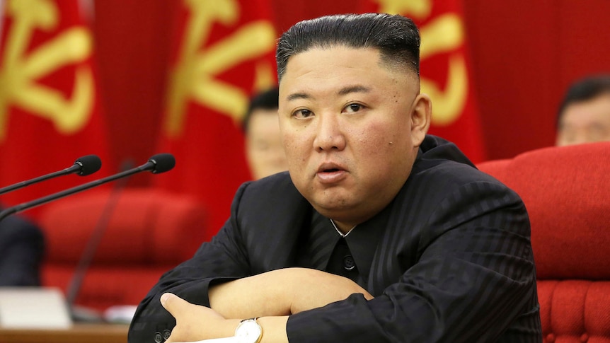 Kim Jong Un sitting at a desk with his arms folded in front of him before a row of North Korean flags