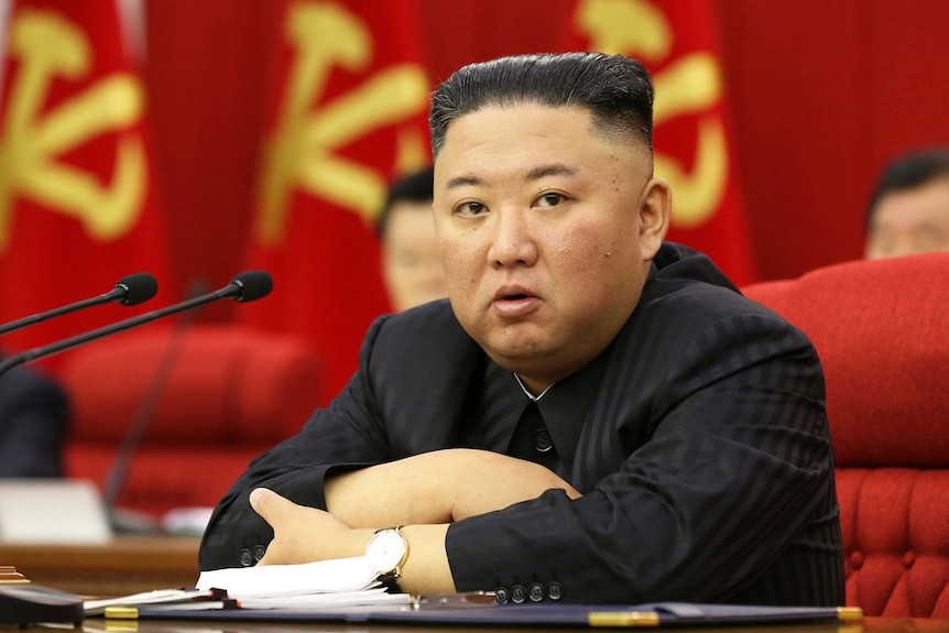 Kim Jong Un sitting at a desk with his arms folded in front of him before a row of North Korean flags