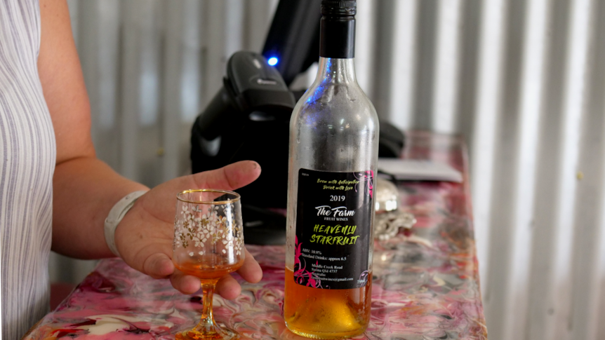 A bottle of yellow fruit wine on a bar with a hand cupping a wine glass. 