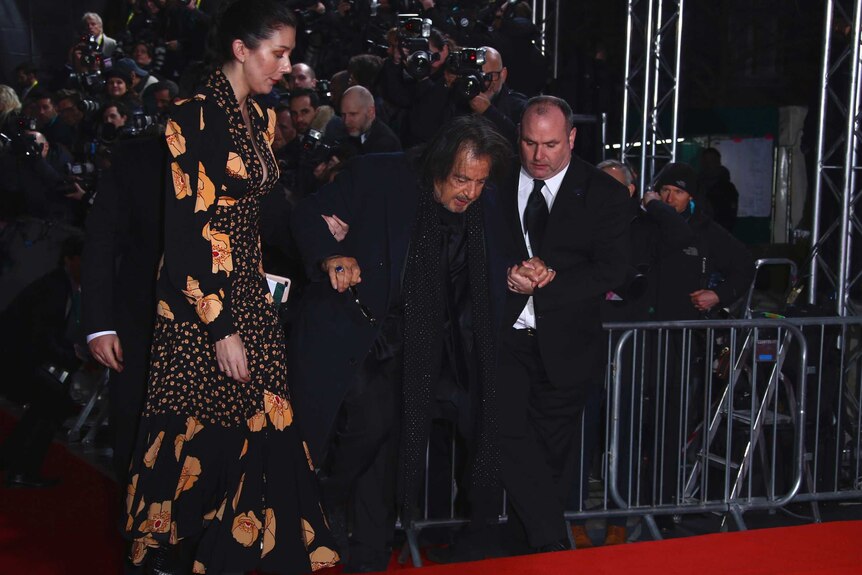 Al Pacino walks up steps covered in red carpet with the help of two men in suits.