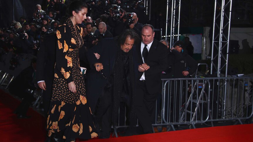 Al Pacino walks up steps covered in red carpet with the help of two men in suits.