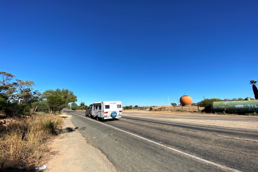 A caravan and car driving on a road with scrub and a big orange sphere on the side.