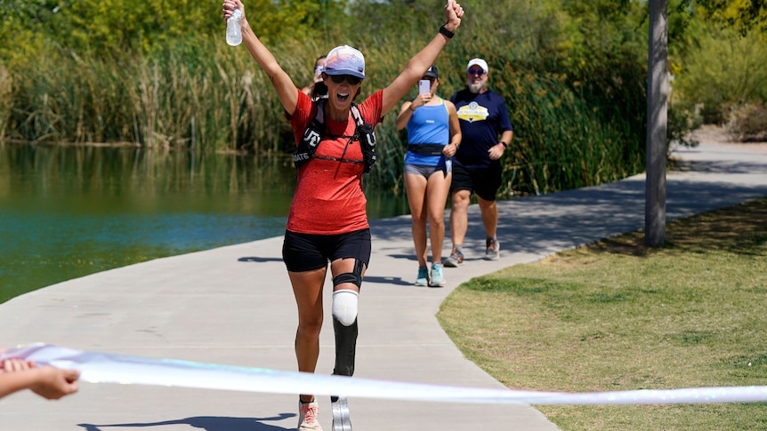 Woman with a prosthetic leg in exercise clothing, cap and sunglasses approaching the finish line with her hands up in the air