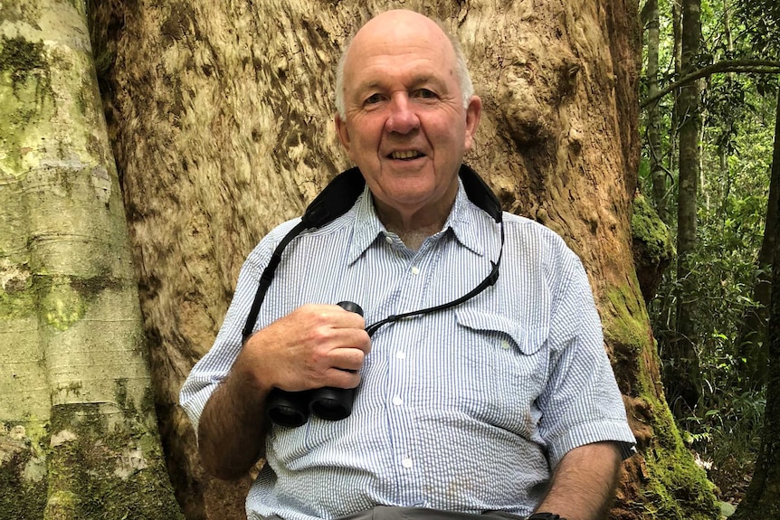 An older man in a linen shirt with a pair of binoculars around his neck smiles at the camera from in front of a tree in a forest