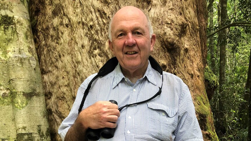 An older man in a linen shirt with a pair of binoculars around his neck smiles at the camera from in front of a tree in a forest