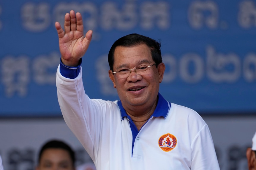 Hun Sen, an older man wearing rimless glasses and a white jumper with blue collar, waves from a stage