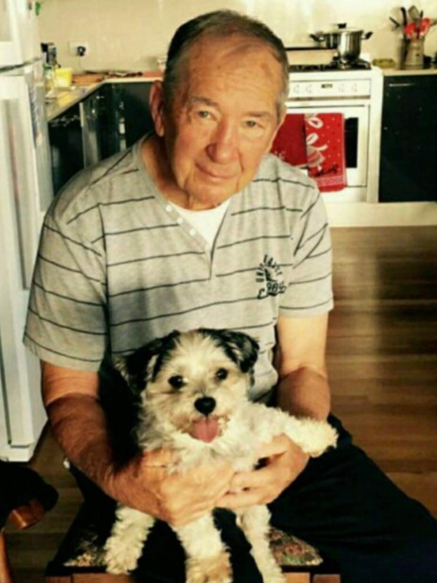 An older man sits in a kitchen holding a small white and black fluffy dog.