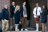 A group of seven teens stand against a brick wall, looking back at the camera. 