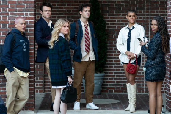 A group of seven teens stand against a brick wall, looking back at the camera. 