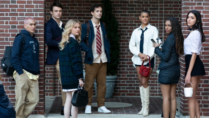 A group of seven teens stand against a brick wall, looking back at the camera. 