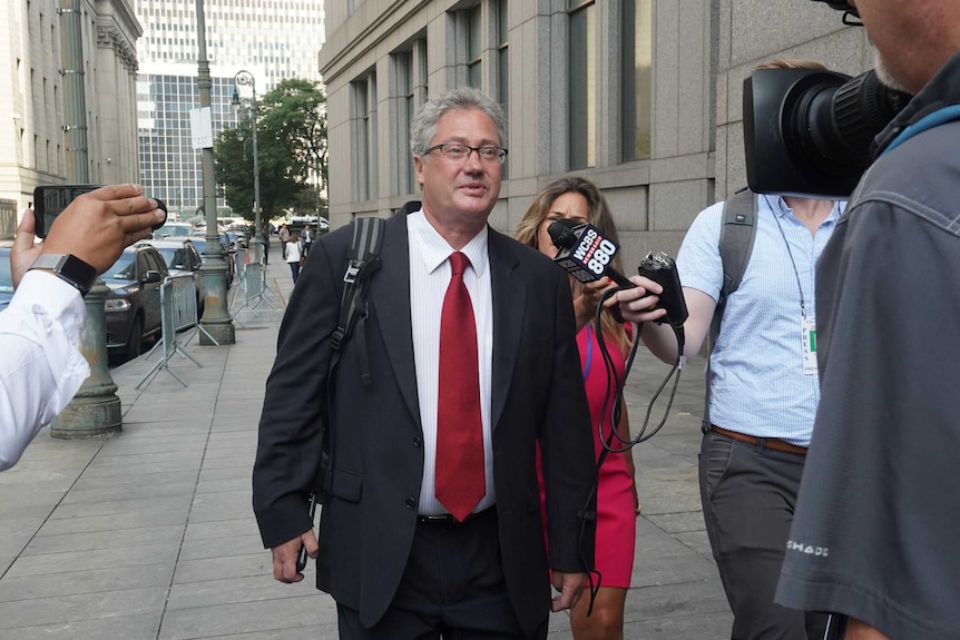 A man in a tie surrounded by reporters walks down a sidewalk