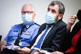 A man speaks through his mask while holding a piece of paper, while sitting down between two people.