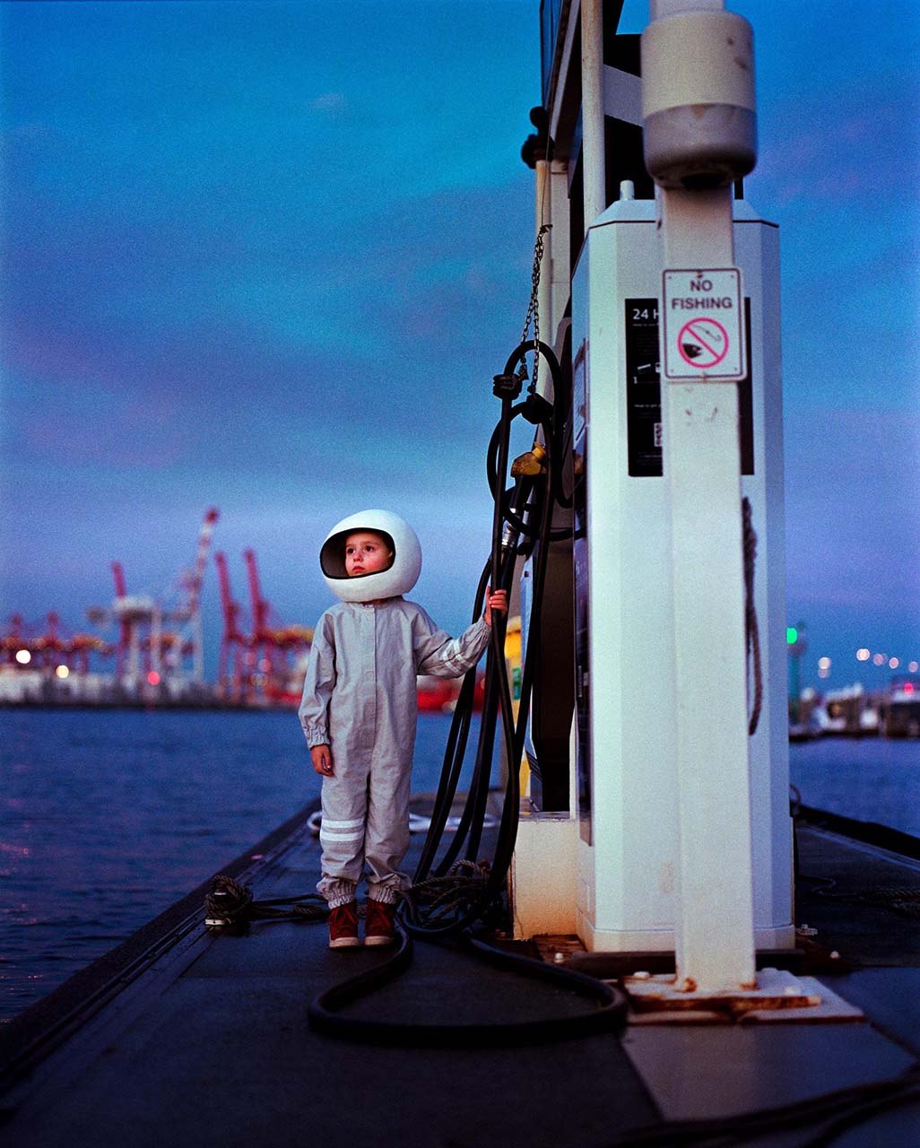 A young girl wearing a spacesuit costume and helmet stands next to a bayside fuel bowser with cranes behind her